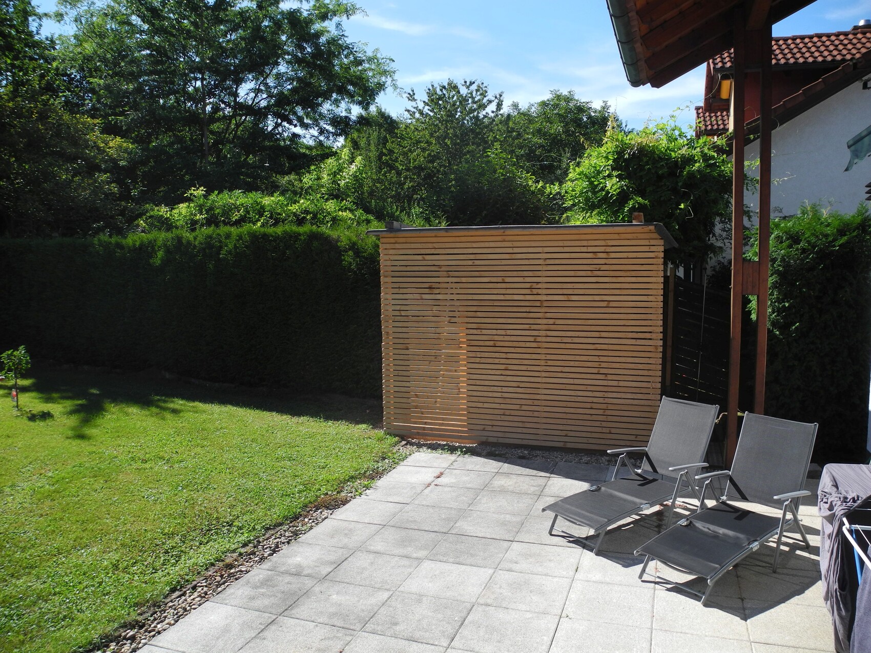 Garten mit Holz-Sichtschutzwand, drei Lounge-Stühle auf grauer Terrasse vor grüner Hecke.
