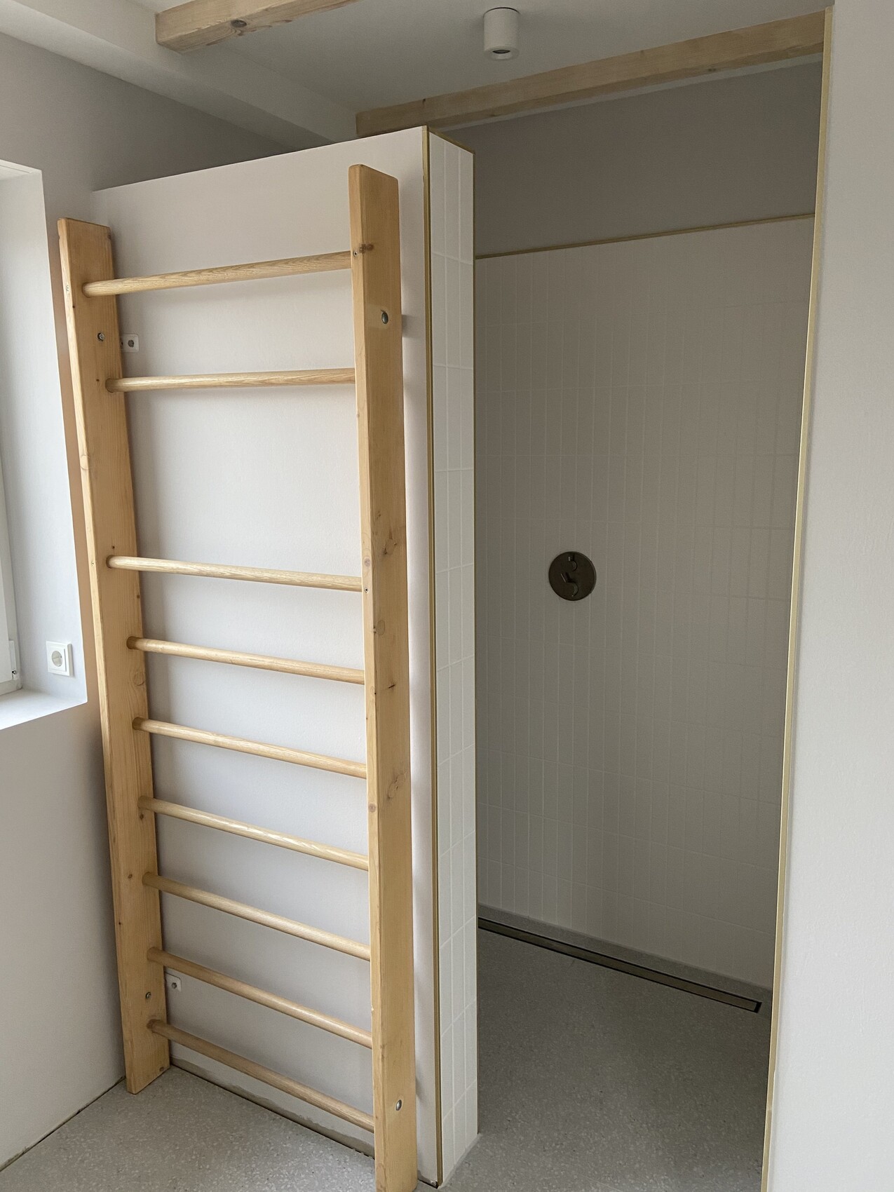 Holz-Sprossenwand steht neben einer Dusche mit weißen Fliesen und runder Armatur.