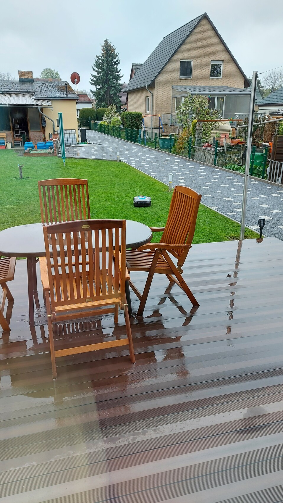 Garten mit regennasser Holzterrasse, runder Esstisch und Holzstühle, Roboter-Rasenmäher auf Gras.