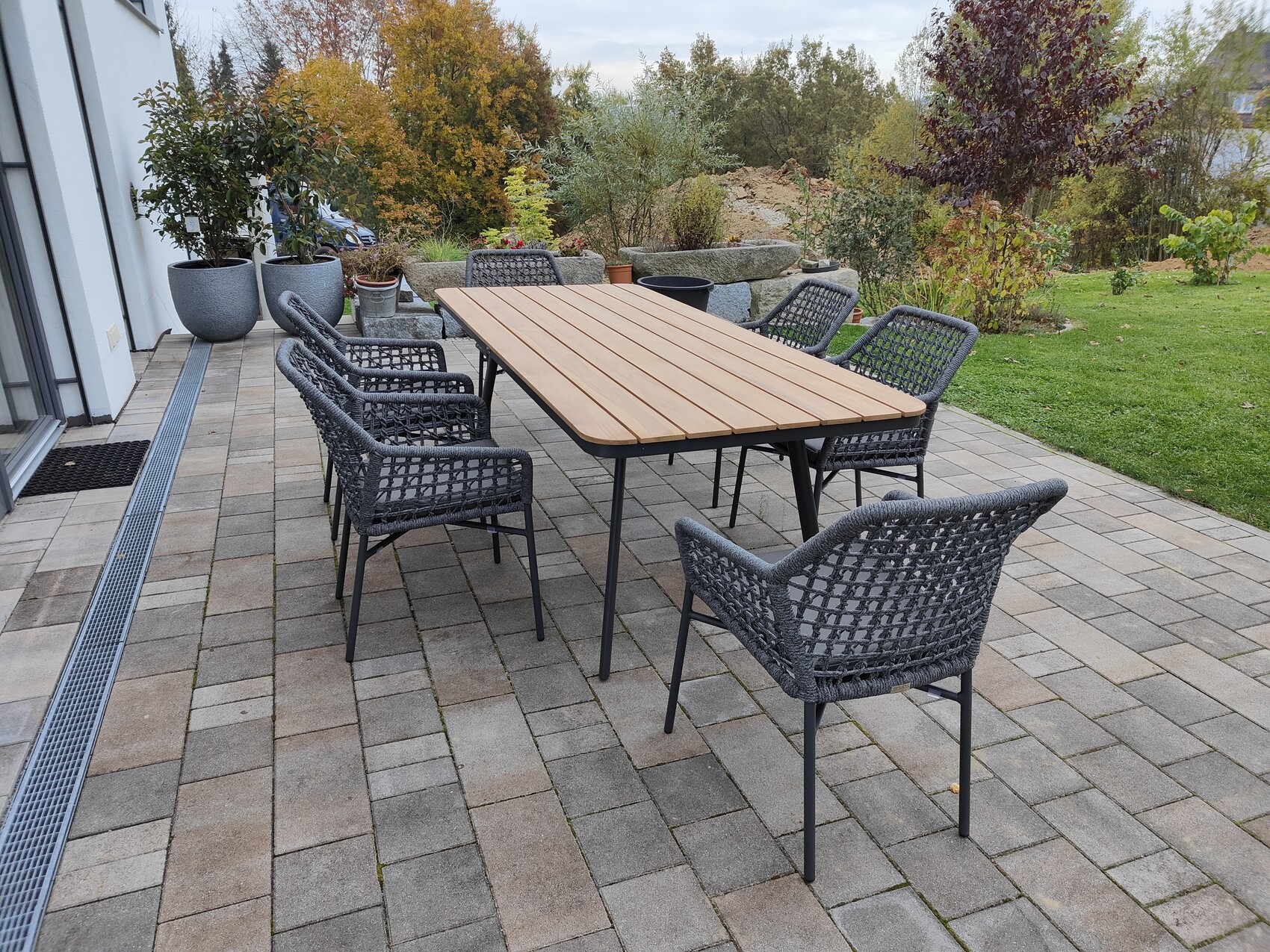 Garten-Terrasse mit langem Holztisch und grauen Geflechtstühlen, von Pflanzen umgeben.