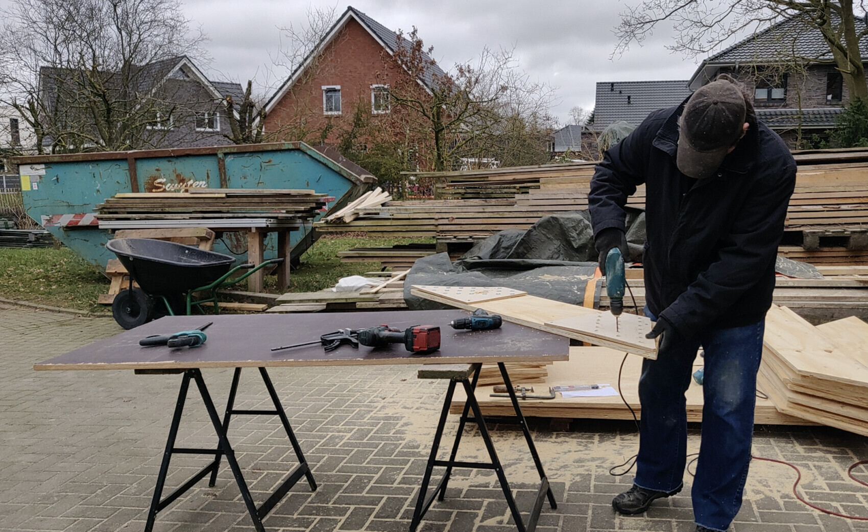 Mann bohrt Löcher in eine Holzplatte im Freien, Holzstapeln und Werkzeuge daneben