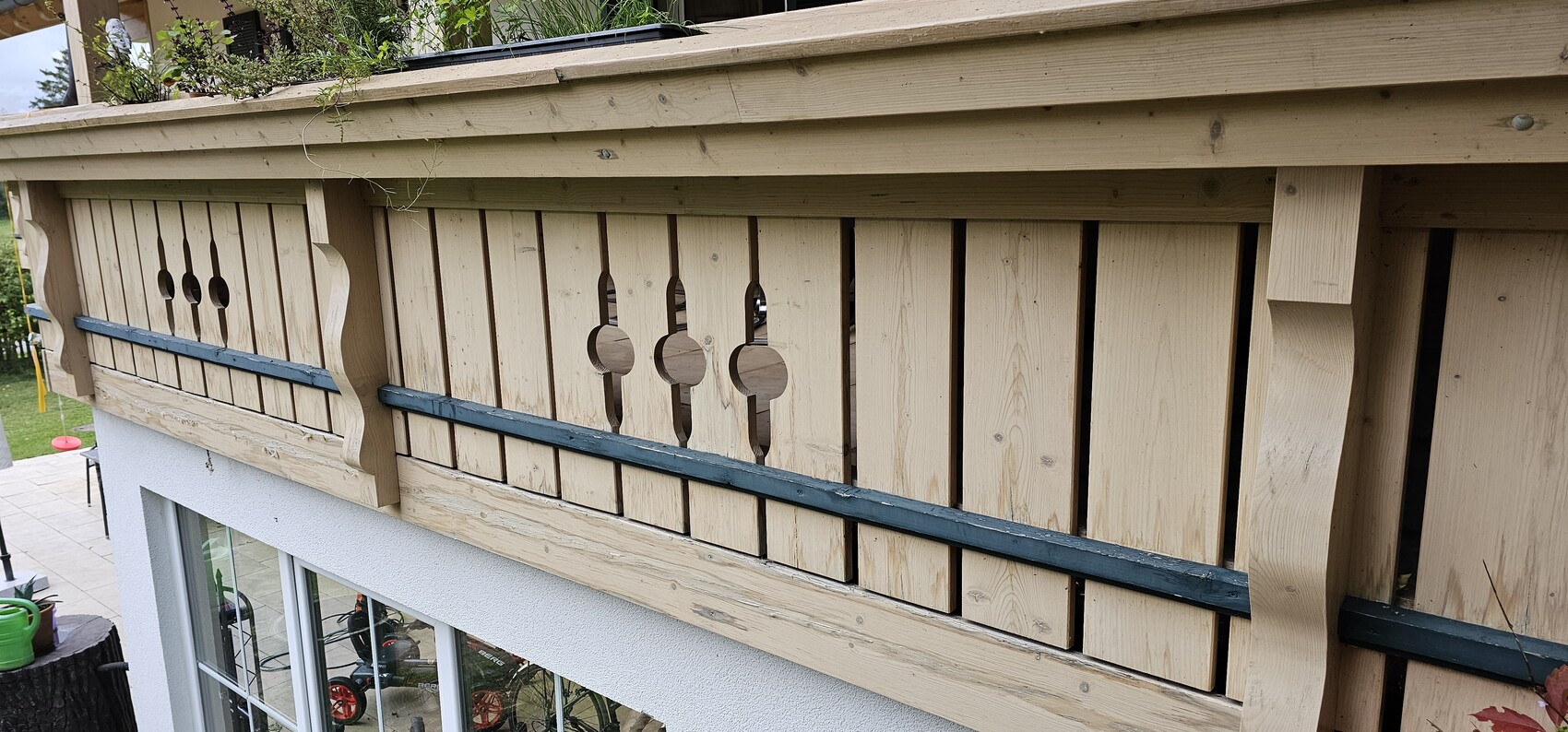 Holzbrüstung eines Balkons mit vertikalen Latten, runden Öffnungen und blauer Zierleiste.