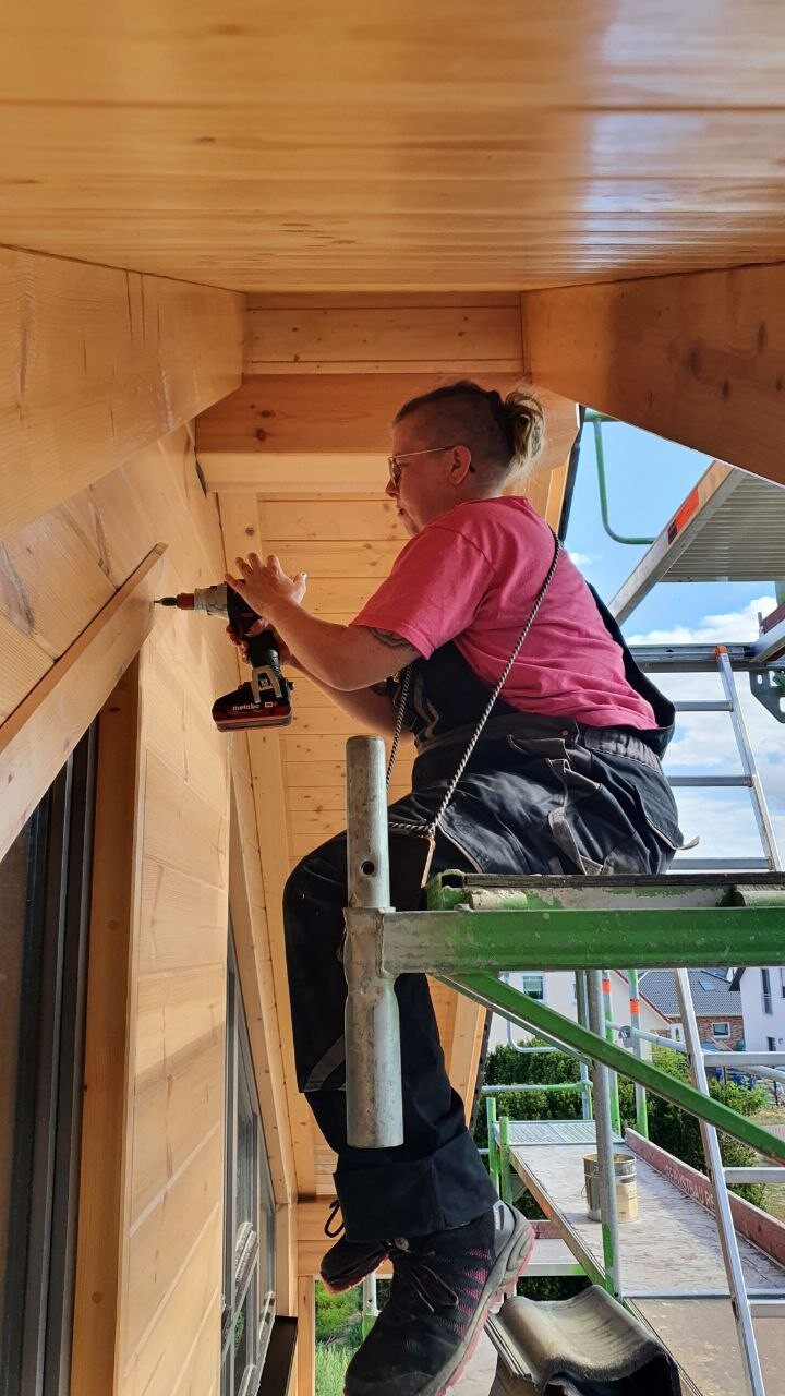 Mann steht auf Baugerüst und bohrt Holzelement in Holzfassade bei Bauarbeiten