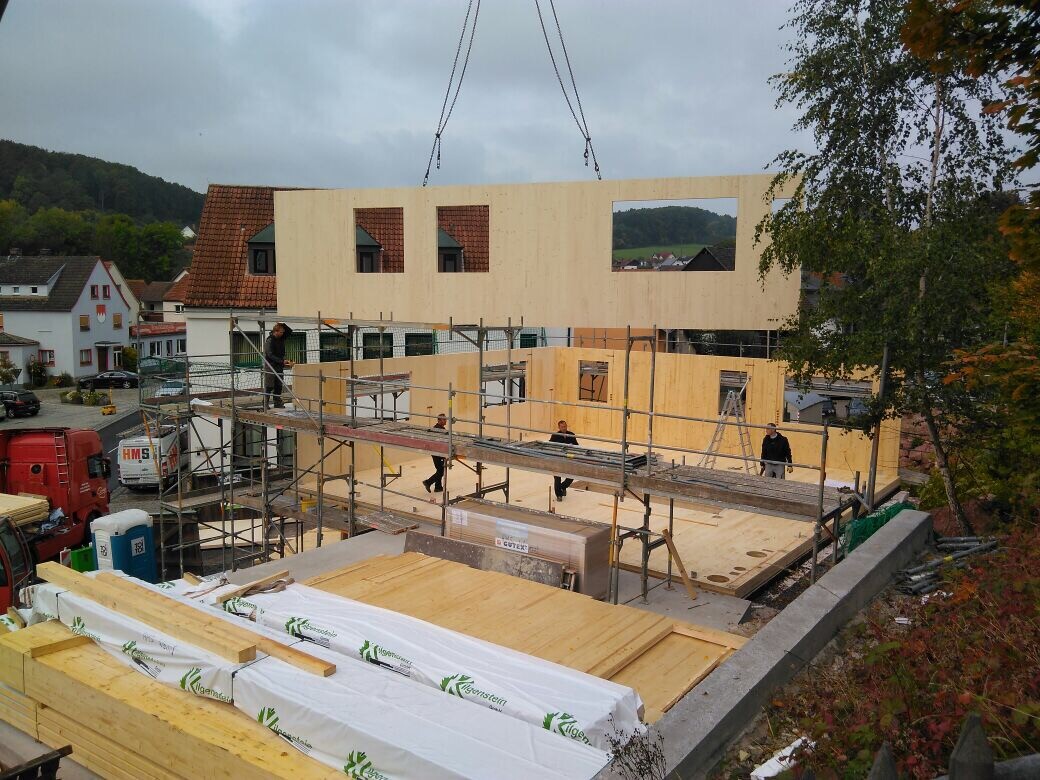 Holzhaus-Bau mit Gerüst: Bauarbeiter montieren Holzwandplatten mit Fensteröffnungen.