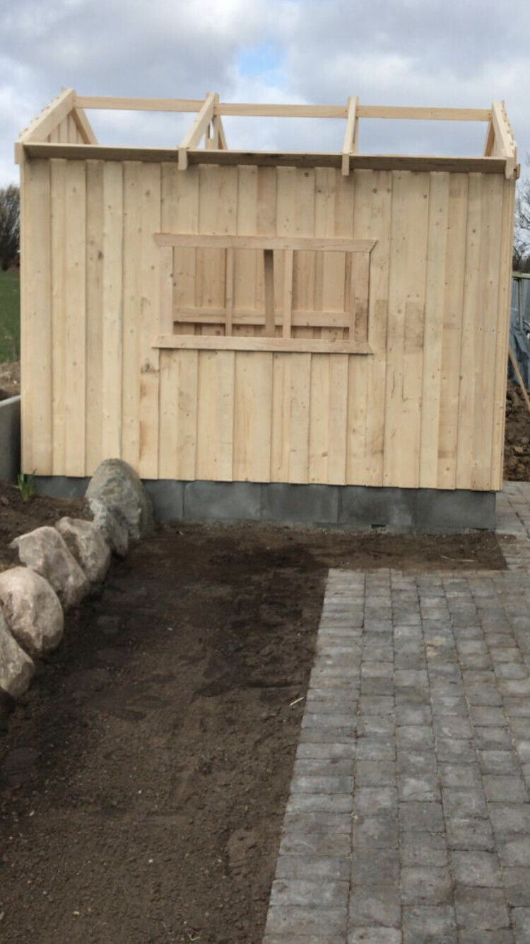 Unfertiges Gartenhaus aus Holzlatten mit offener Fensteröffnung auf Betonfundament.