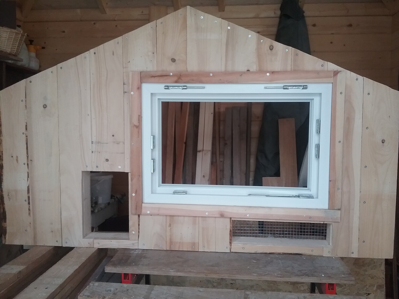 Holzschuppen mit schrägem Dach, weißem Fensterrahmen, kleine linke Öffnung und Lüftungsgitter unten.