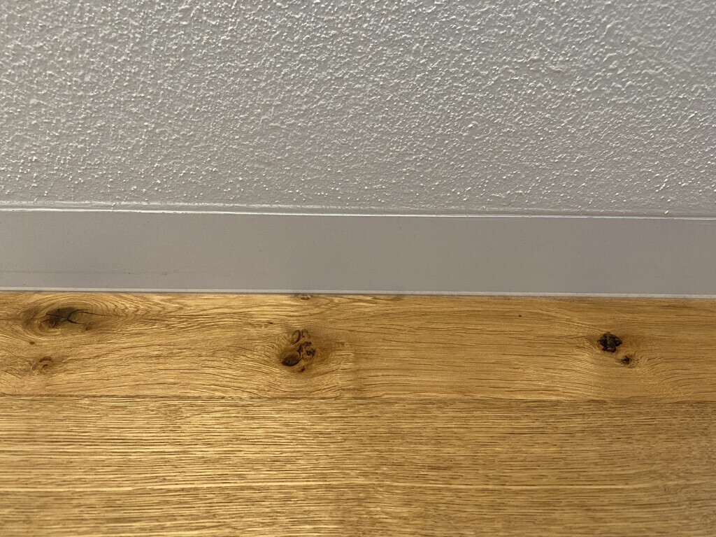 Texturierte weiße Wand über grauer Sockelleiste, Holzfußboden mit Knoten.