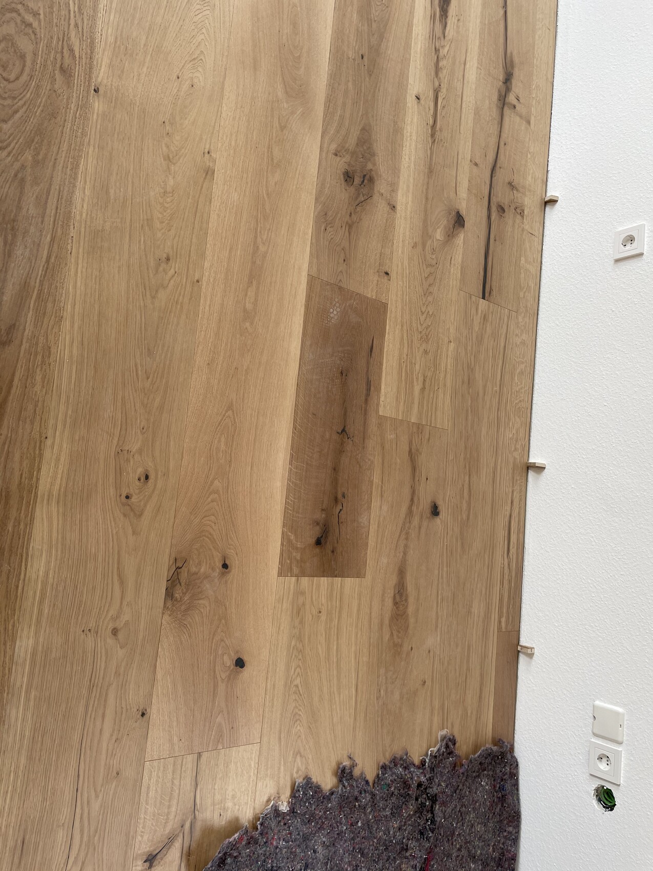 Heller Eichendielen-Holzboden neben weißer Wand mit Steckdosen; untere Kante mit Teppichrest.