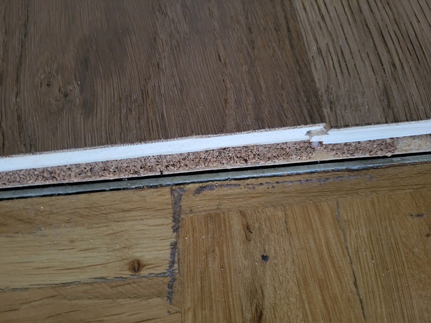 Nahaufnahme eines Holzfußbodens mit weißer Leiste; darunter Kork-Unterlage und eine Spalte zwischen den Dielen.