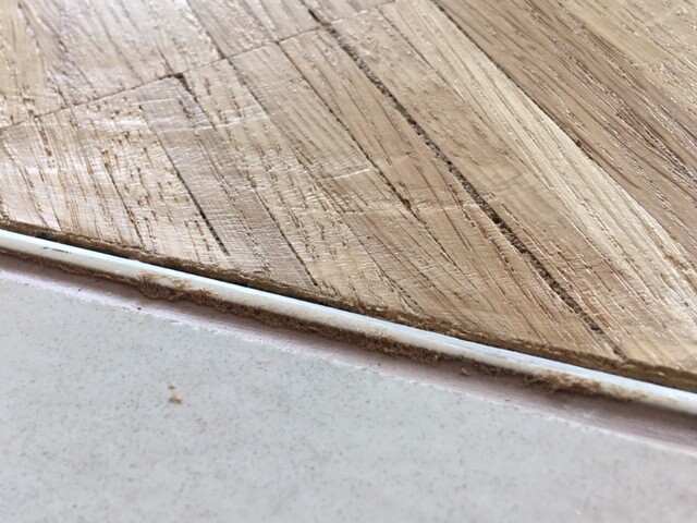 Nahaufnahme von Holzboden mit Maserung, daneben graue glatte Fläche und Fuge.