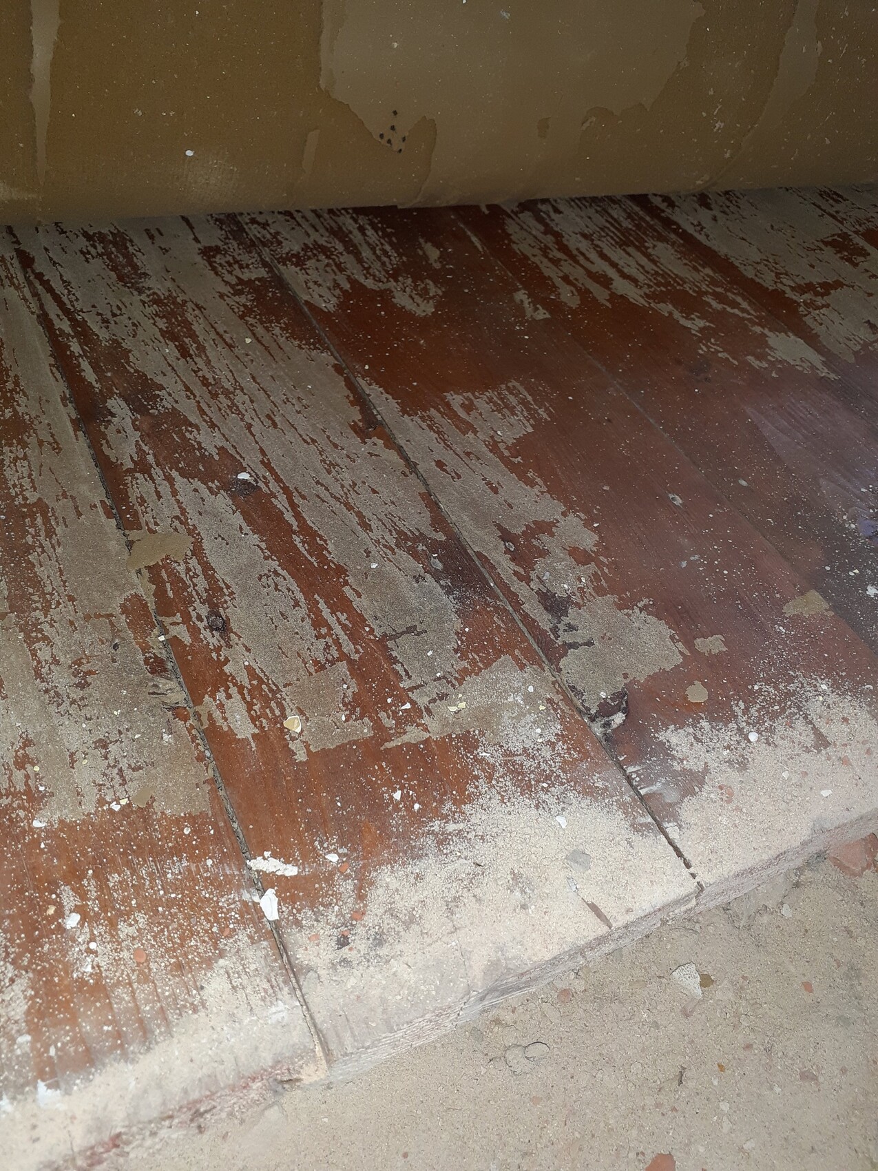 Abgenutzter Holzboden mit abblätterndem Lack, Staub und Schmutz; Wand mit Putzabplatzungen.