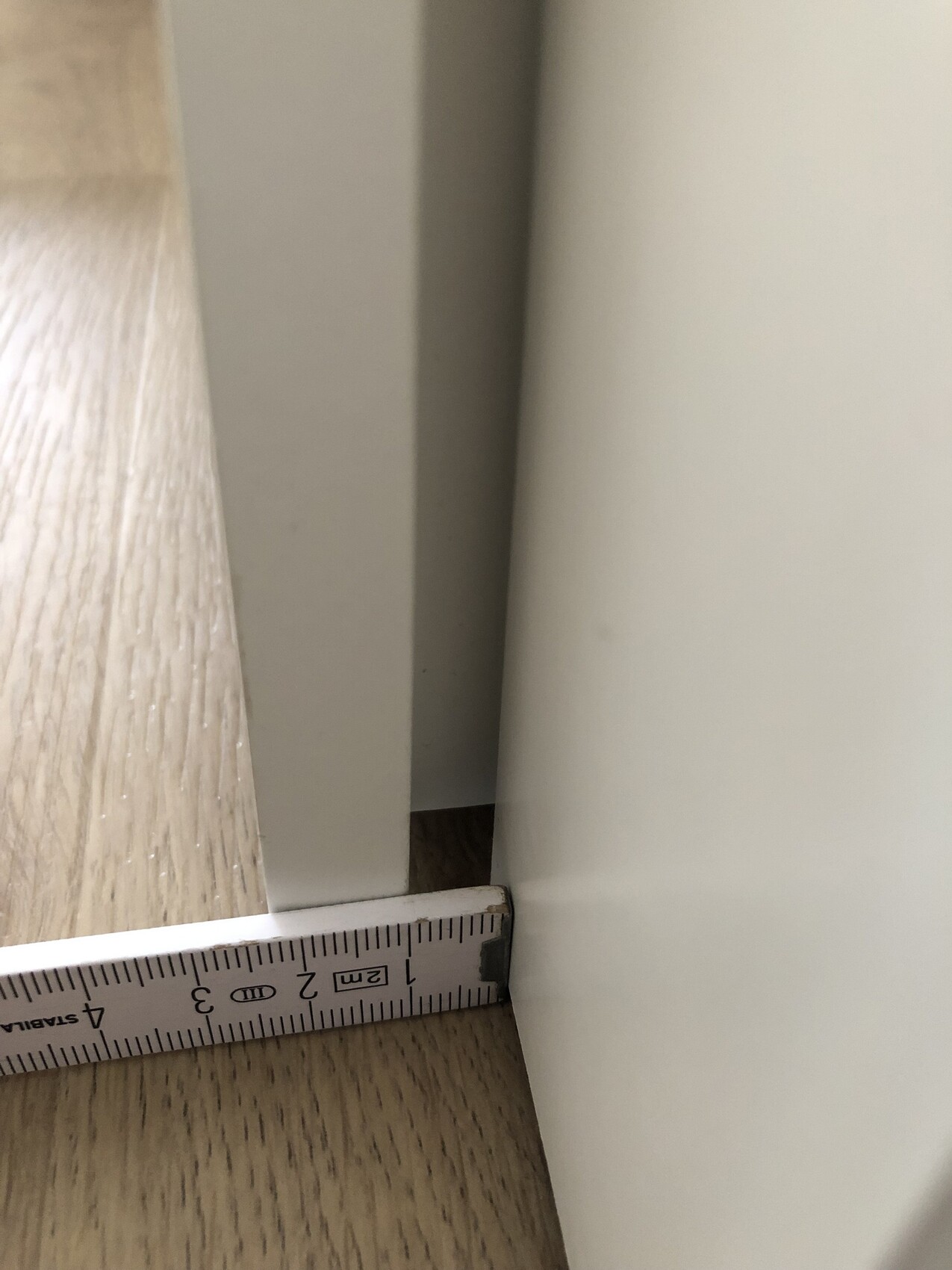 Nahaufnahme: Holzfußboden links, graue Kante, weißes Möbelteil rechts; Maßband zeigt ca. 2 cm Spalt.