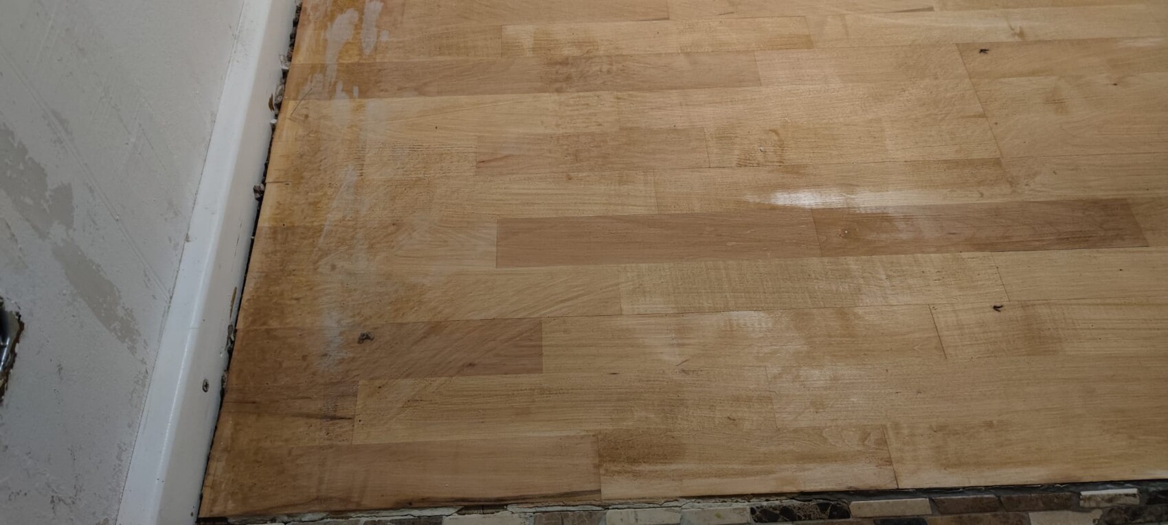 Holzboden mit Flecken und Staub, Randbereich neben weißer Wand und Sockelleiste.