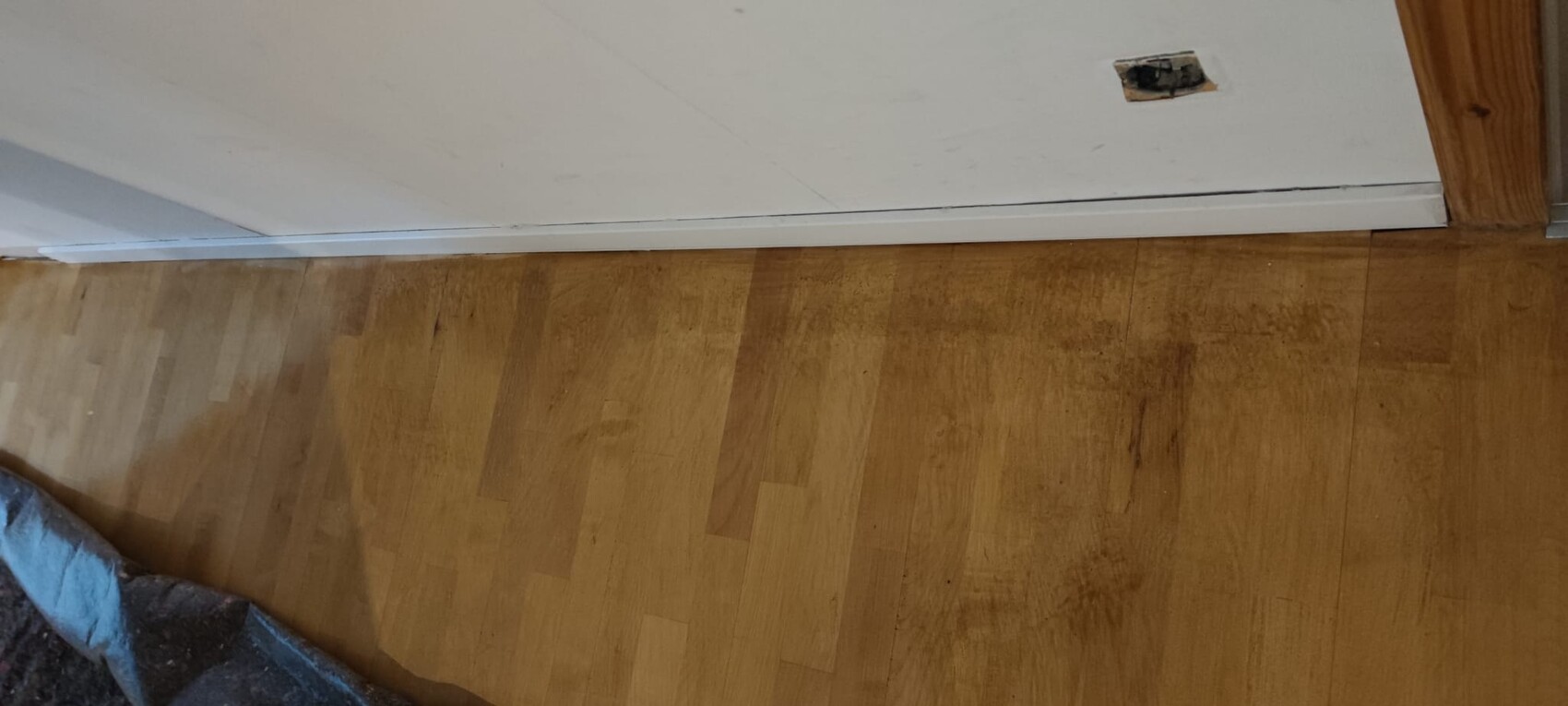 Heller Holzboden mit weißen Sockelleisten; kleines rechteckiges Loch in der Wand.