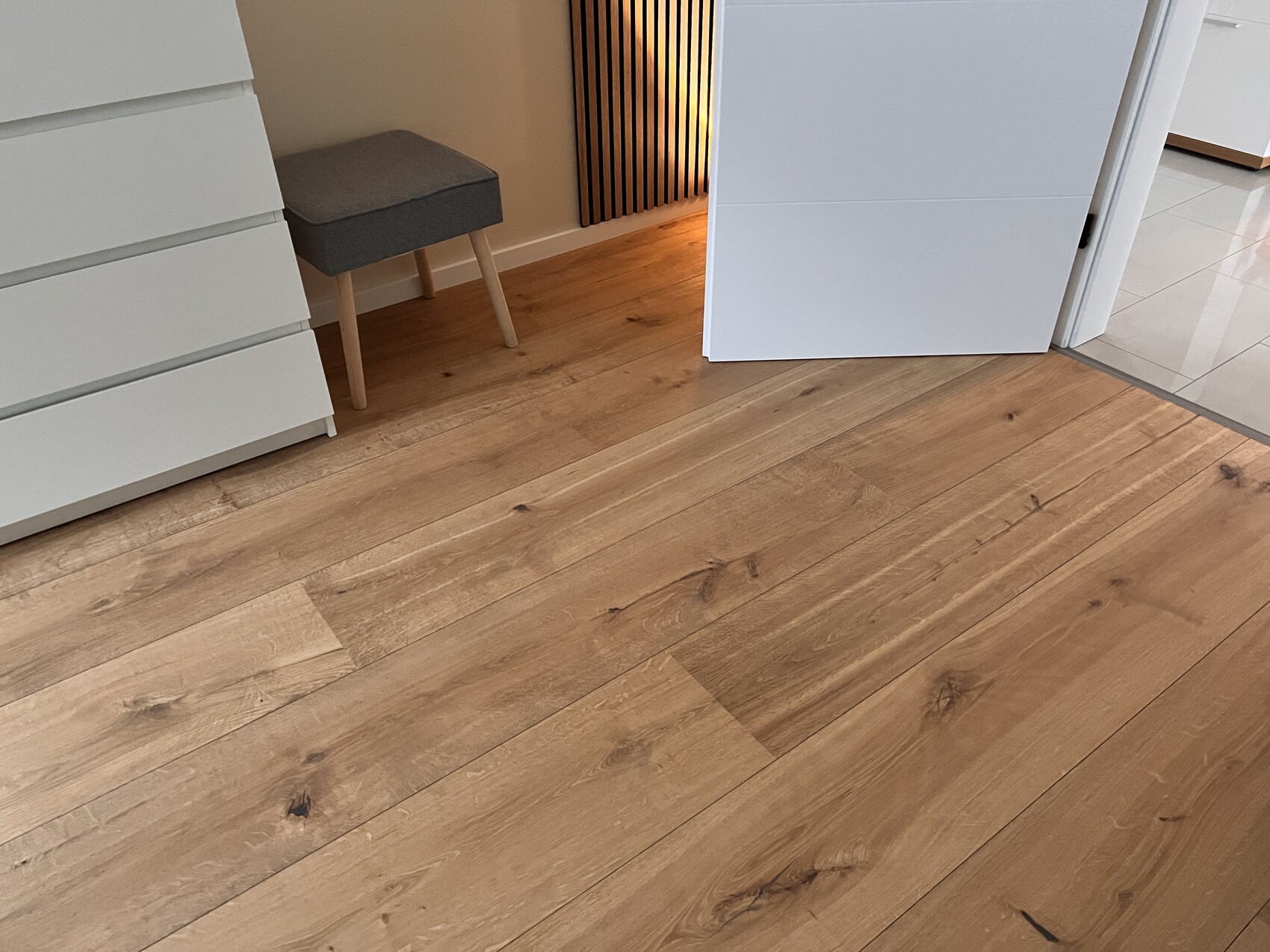 Weiße Möbel, grauer Hocker, offener Schrank rechts, warmer Lichtstrahl hinter Holzboden.