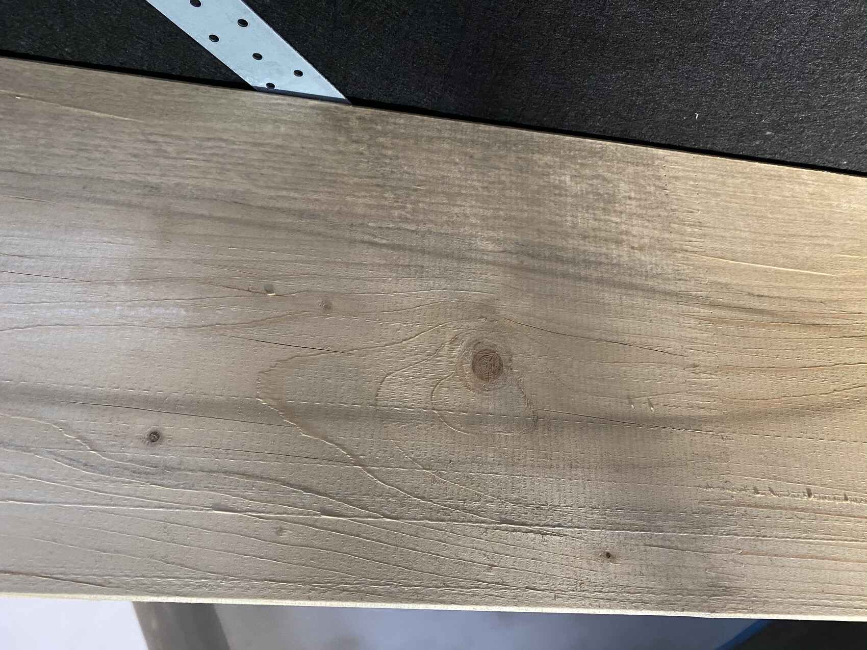 Holzplatte mit sichtbarer Maserung und Knoten; oben links ein perforiertes Metallwinkelstück auf dunklem Hintergrund.