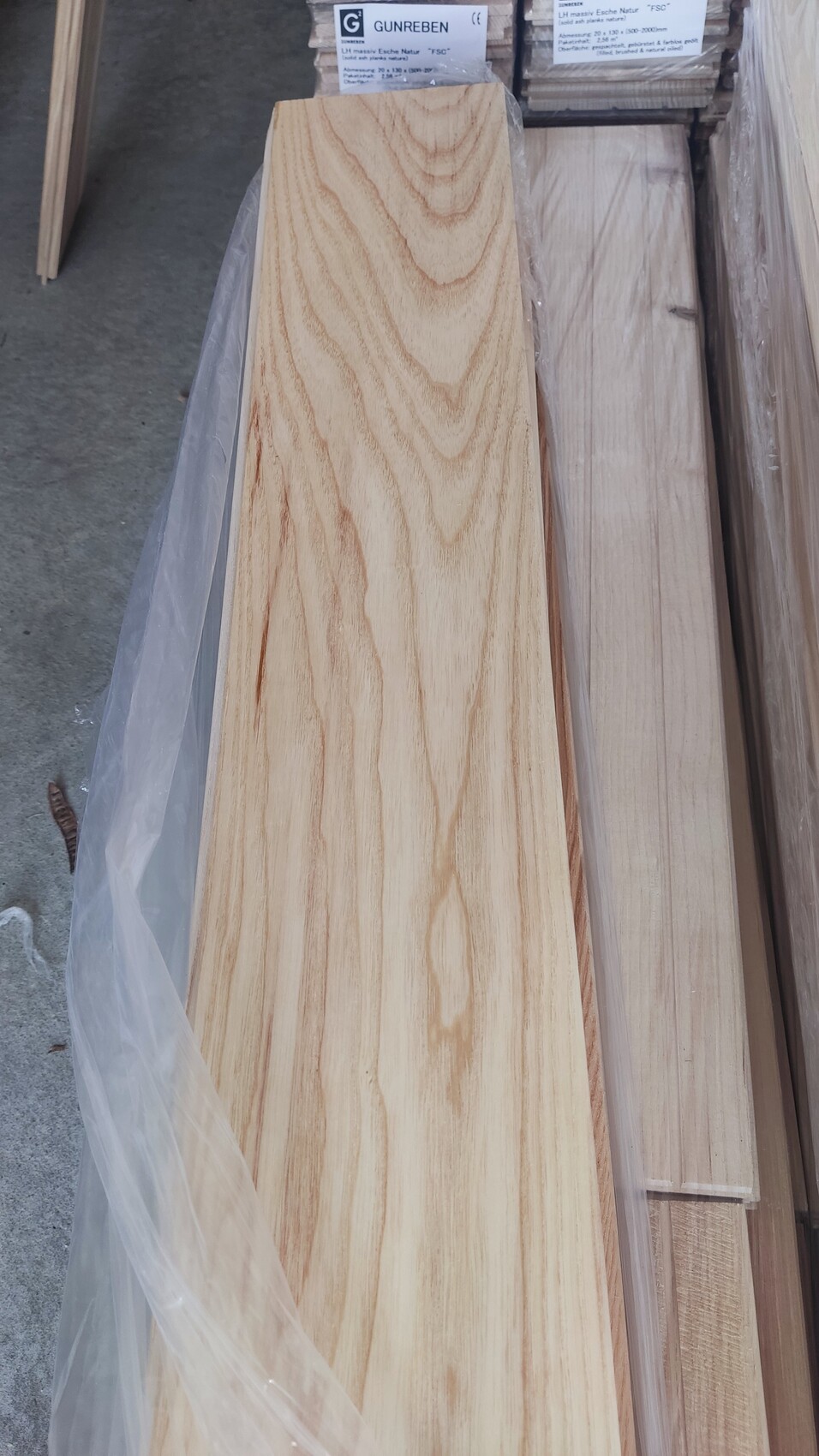 Längliches Holzbrett mit heller Maserung, umgeben von weiteren Holzbrettern in Plastikfolie.