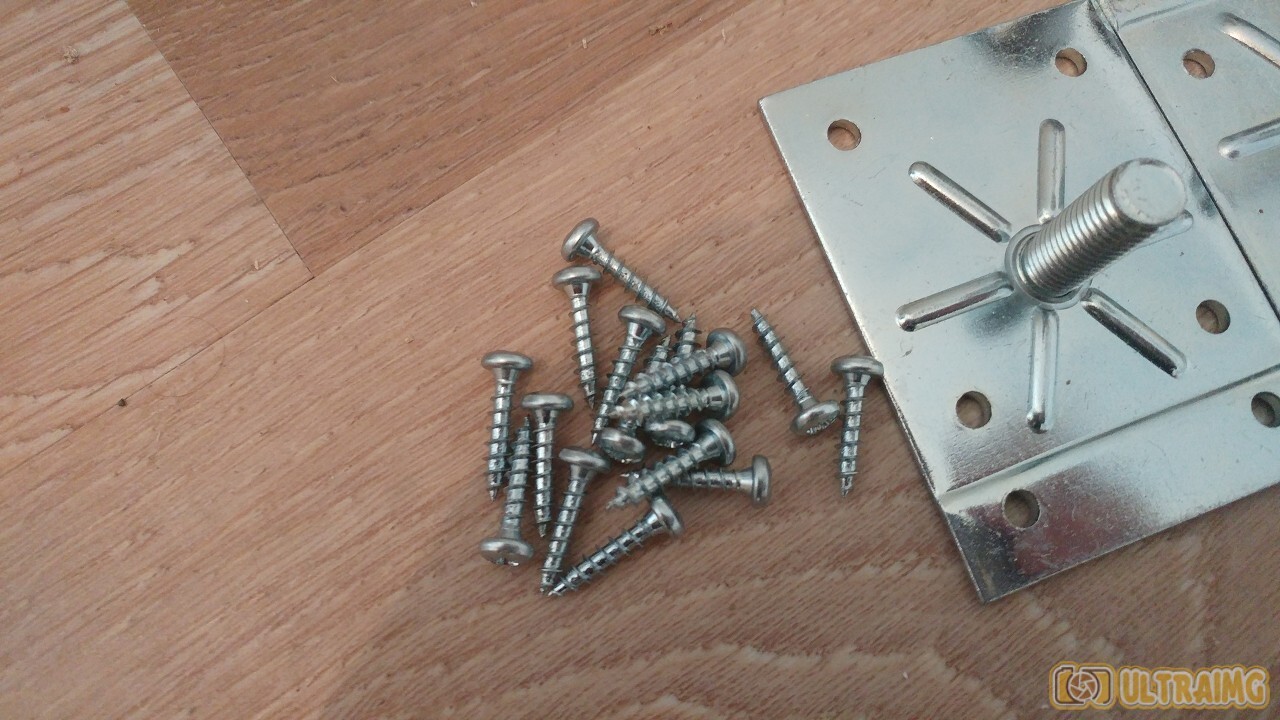 Mehrere Schrauben liegen auf Holzboden neben einer silbernen Metallplatte mit zentralem Gewinde.