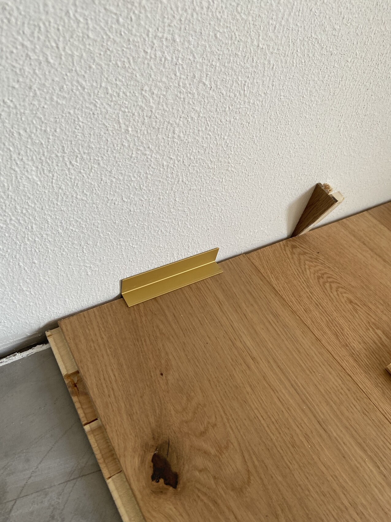 Holzplatte liegt auf dem Boden vor einer weißen, texturierten Wand; goldene Metallleiste ruht darauf