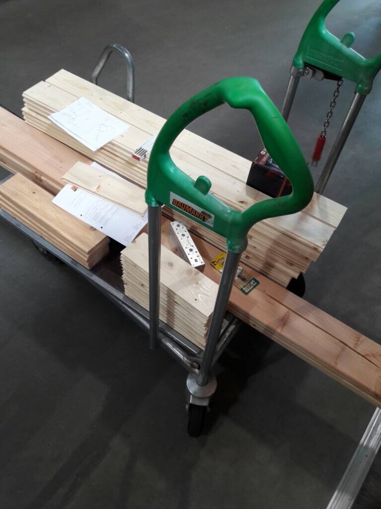 Handwagen mit Holzstapeln in einer Bauwerkstatt, grüne Griffe deutlich sichtbar.