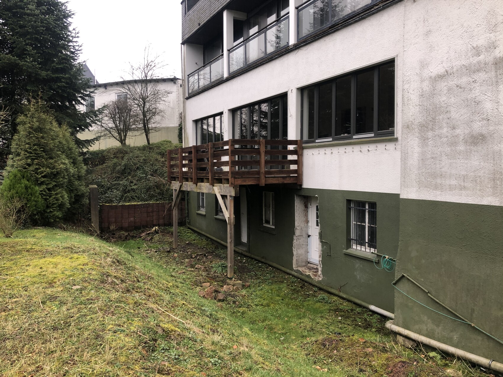 Wohnhaus mit einer hölzernen Balkonbrücke über dem Keller, grünem Sockel und Garten.