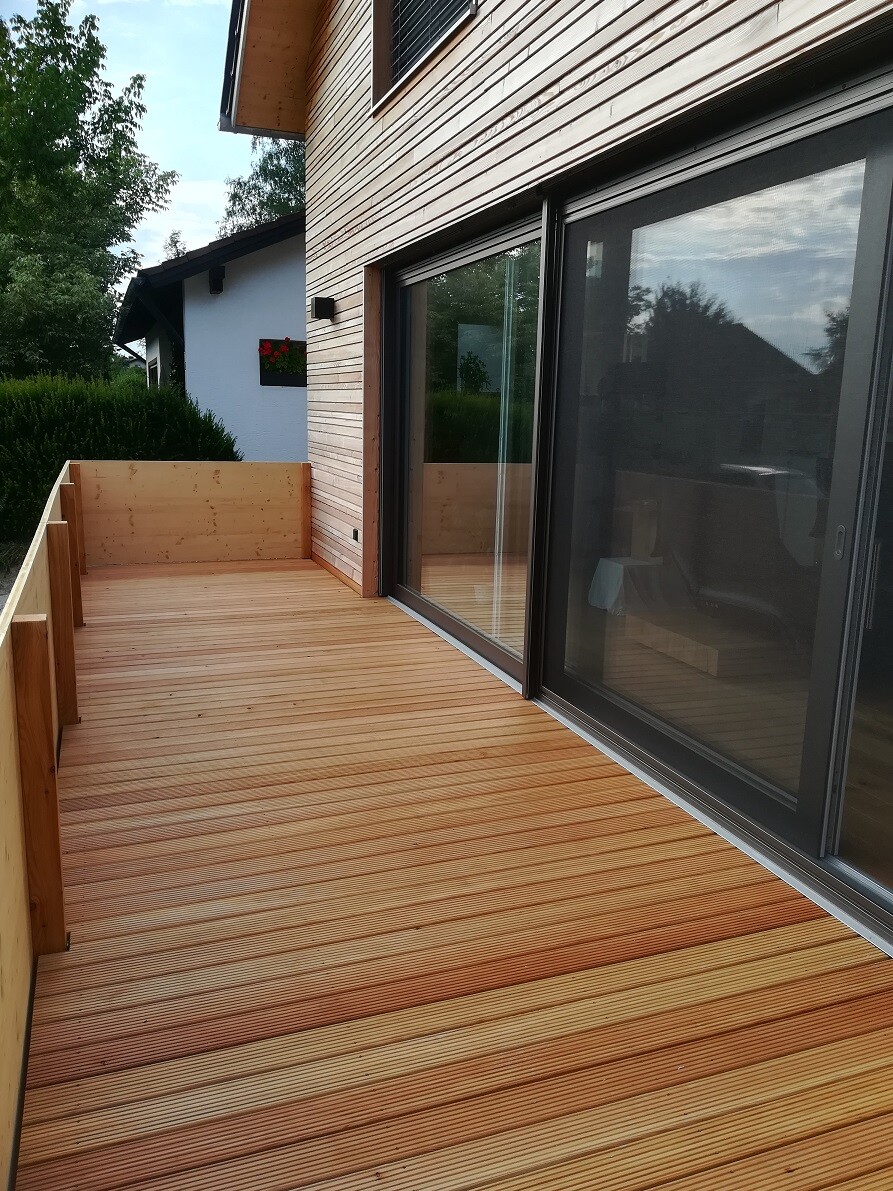 Holzdeck-Terrasse am Haus mit großen Glas-Schiebetüren und Blick auf Garten.