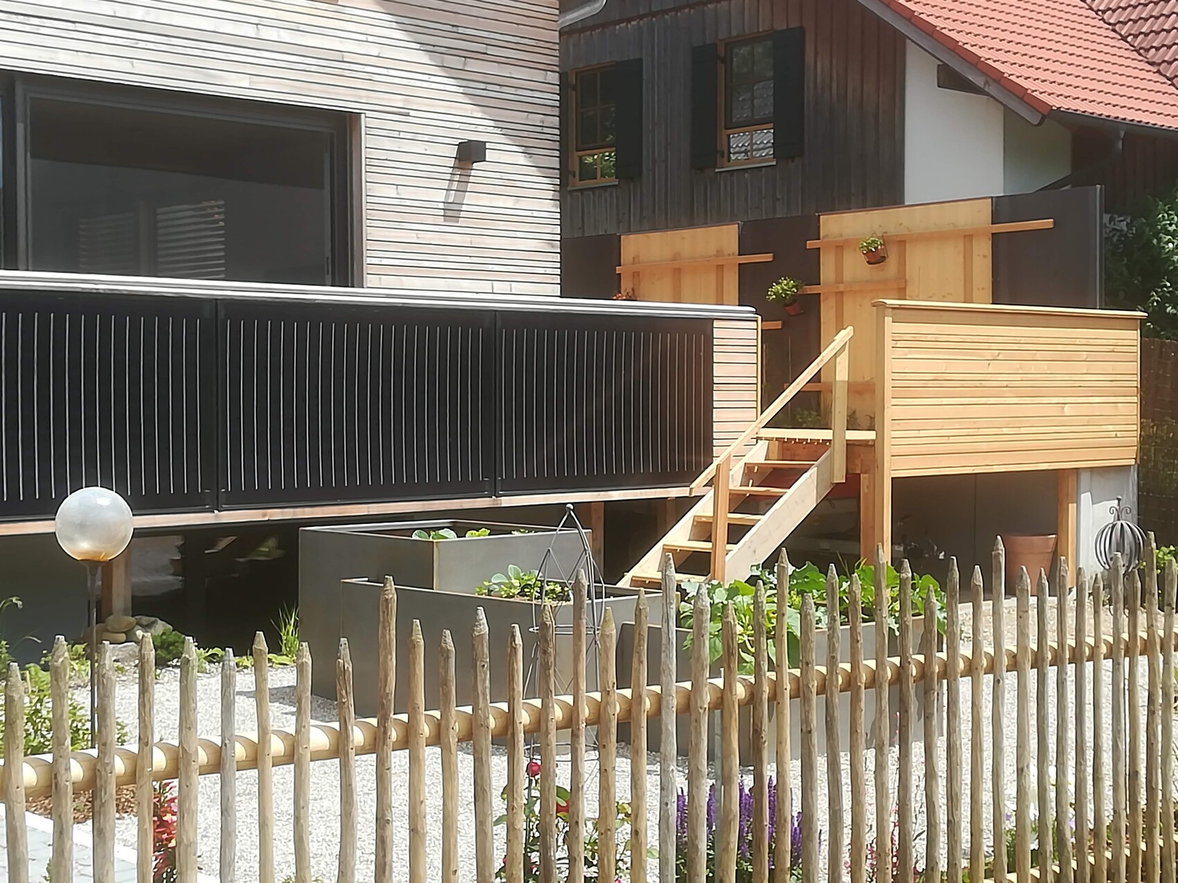 Außenansicht eines Hauses mit schwarzem Geländer, Holzdeck, Treppe und erhöhtem Holzhäuschen im Garten.