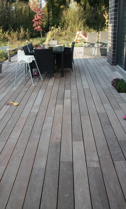Holzterrasse im Freien mit Esstischgruppe aus Rattan und Garten im Hintergrund.