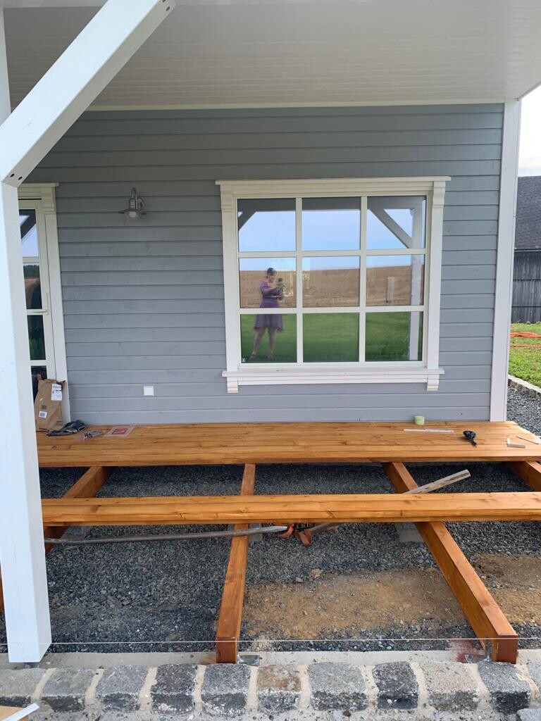 Holzdeck-Unterbau vor blauem Haus; Fenster spiegelt eine Person.