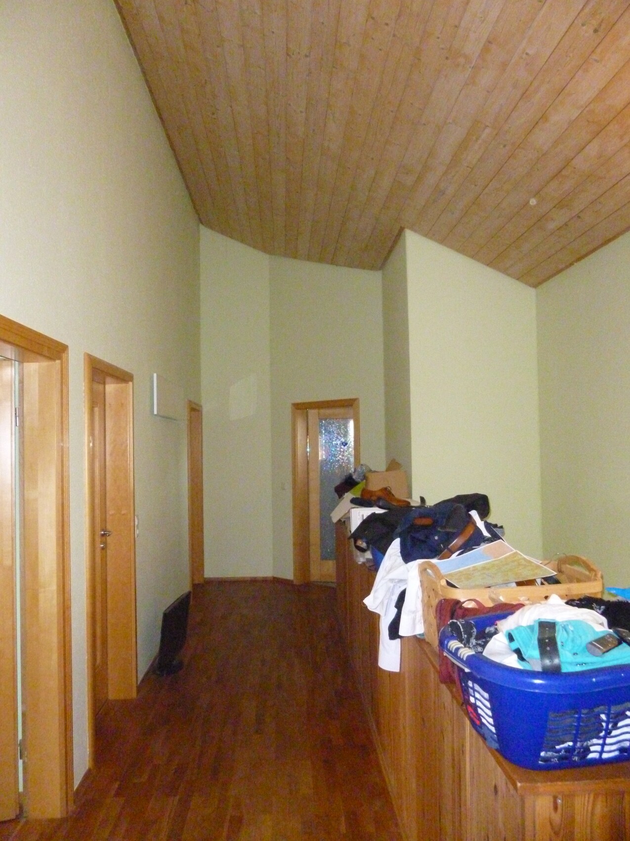Langer Innenflur mit Holzboden und Holzdecke; rechts Kleidung auf Kommode, blauer Wäschekorb.