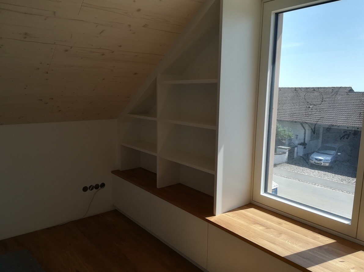 Heller Raum mit schrägem Holzdach, weißen Regalen und Holzfensterbank am Fenster.