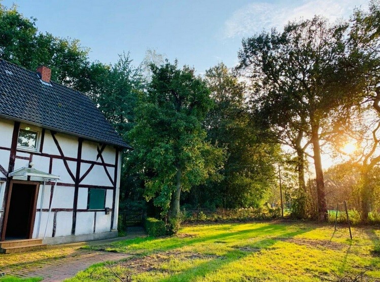 Fachwerkhaus links mit offener Tür, Garten, Bäume und Sonnenuntergang im Hintergrund.