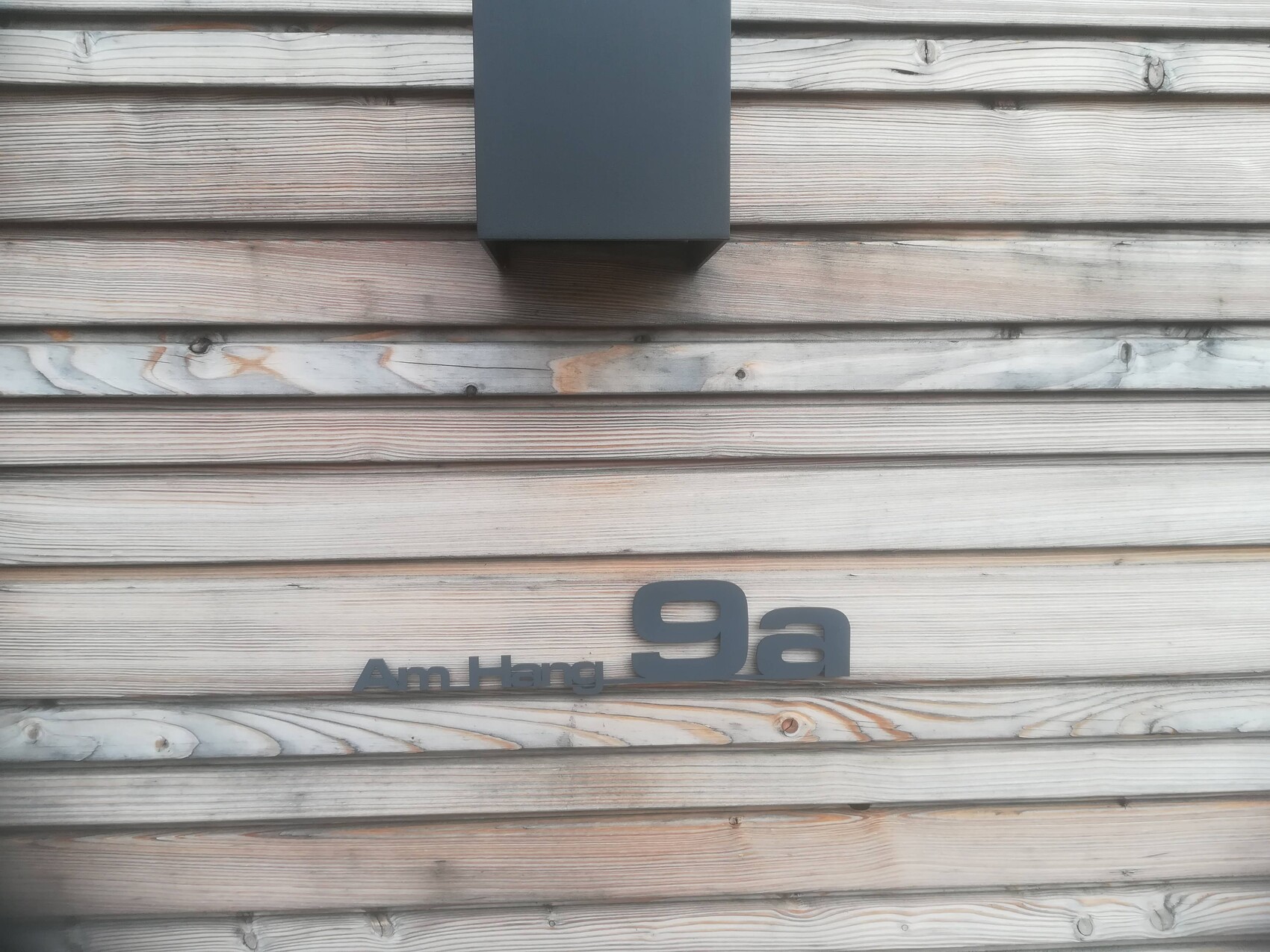 Holzverkleidung einer Hausfassade mit der Aufschrift Am Hang 9a und graue Außenleuchte darüber