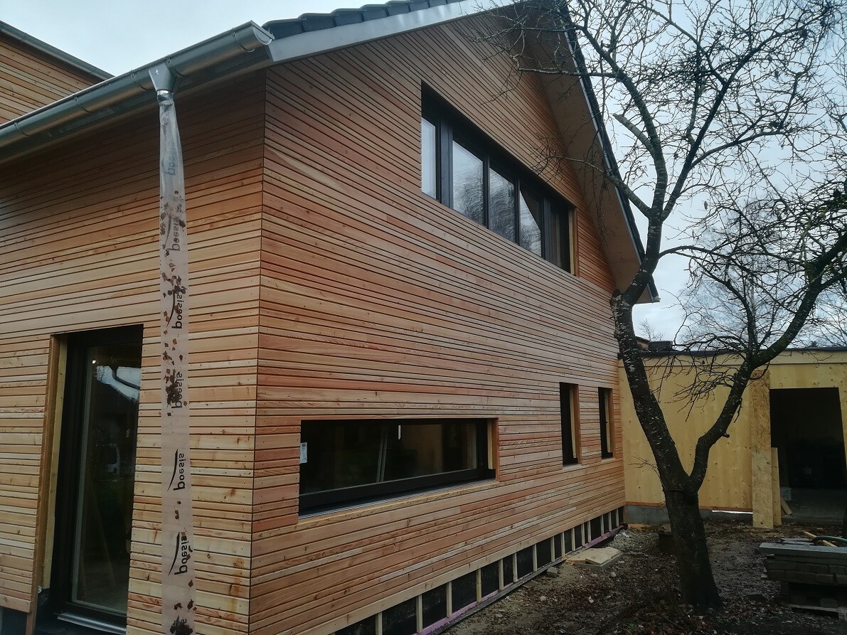 Zweistöckiges Haus mit horizontaler Holzverkleidung, großen Fenstern und Baum im Vordergrund