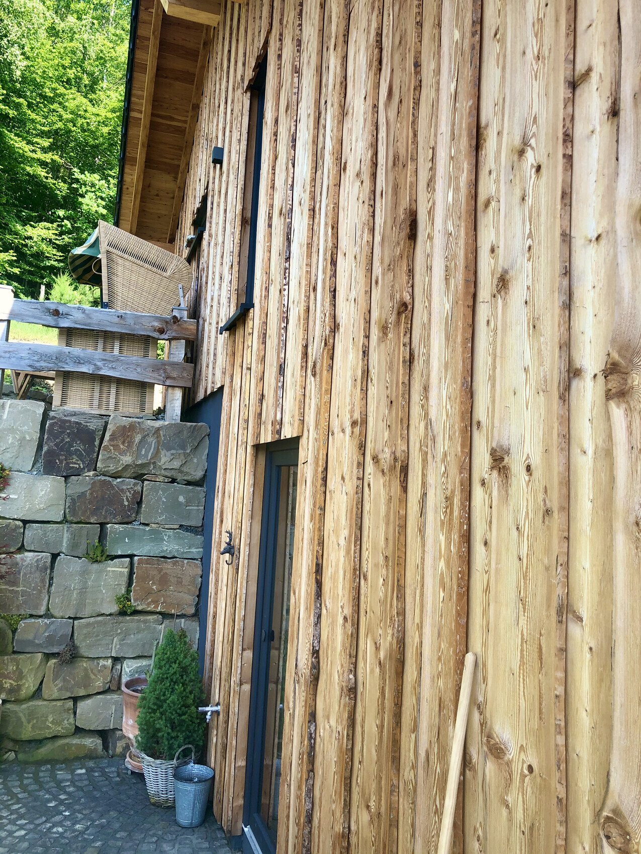 Außenansicht eines Holzhauses mit vertikalen Brettern, blauem Türrahmen, Steinmauer und Pflanzen