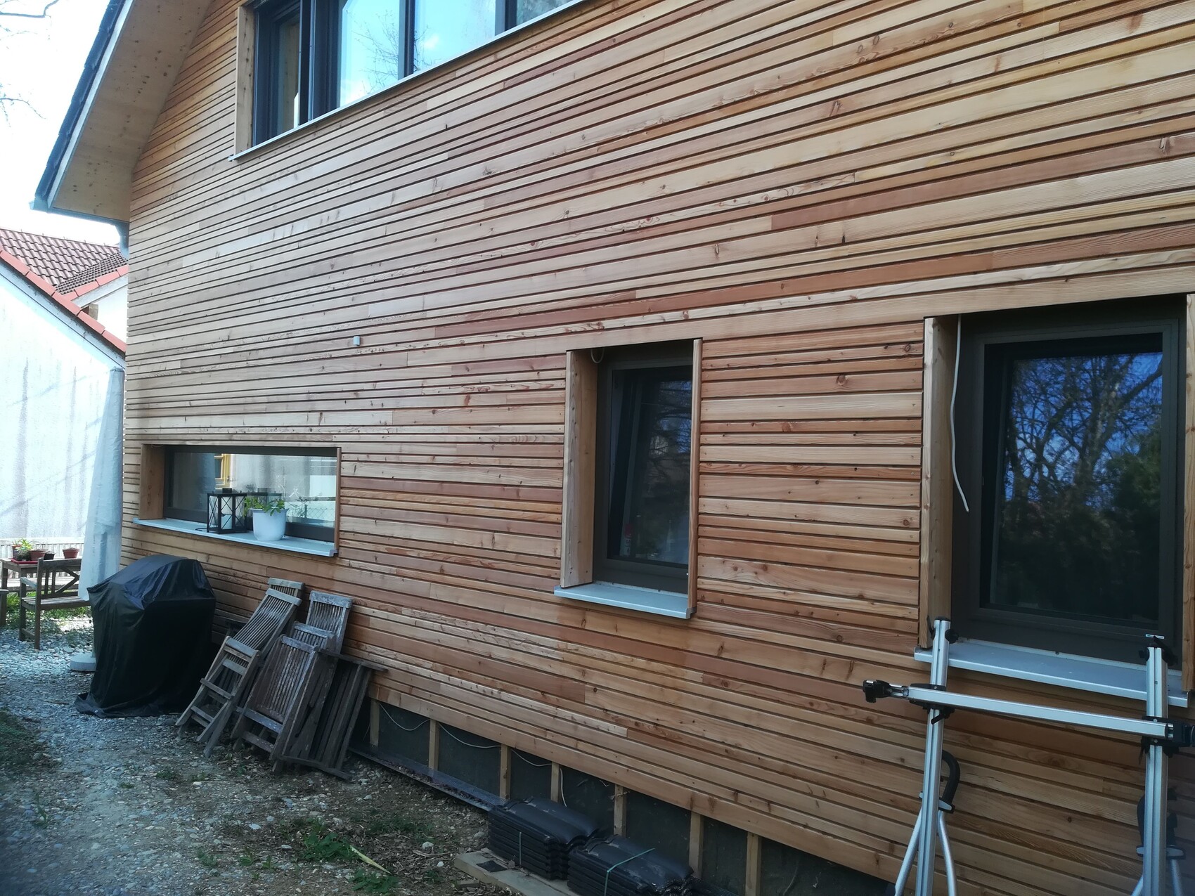 Außenansicht eines Hauses mit horizontaler Holzverkleidung, Fenstern, Grillabdeckung und Leitern.