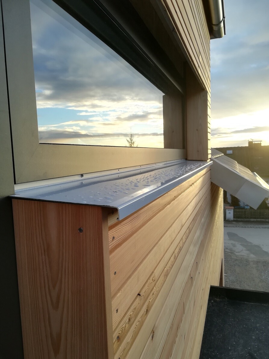 Außenansicht eines Holzhauses mit Fensterfront, schmaler Metallrinne und Wassertropfen, Sonnenuntergang.