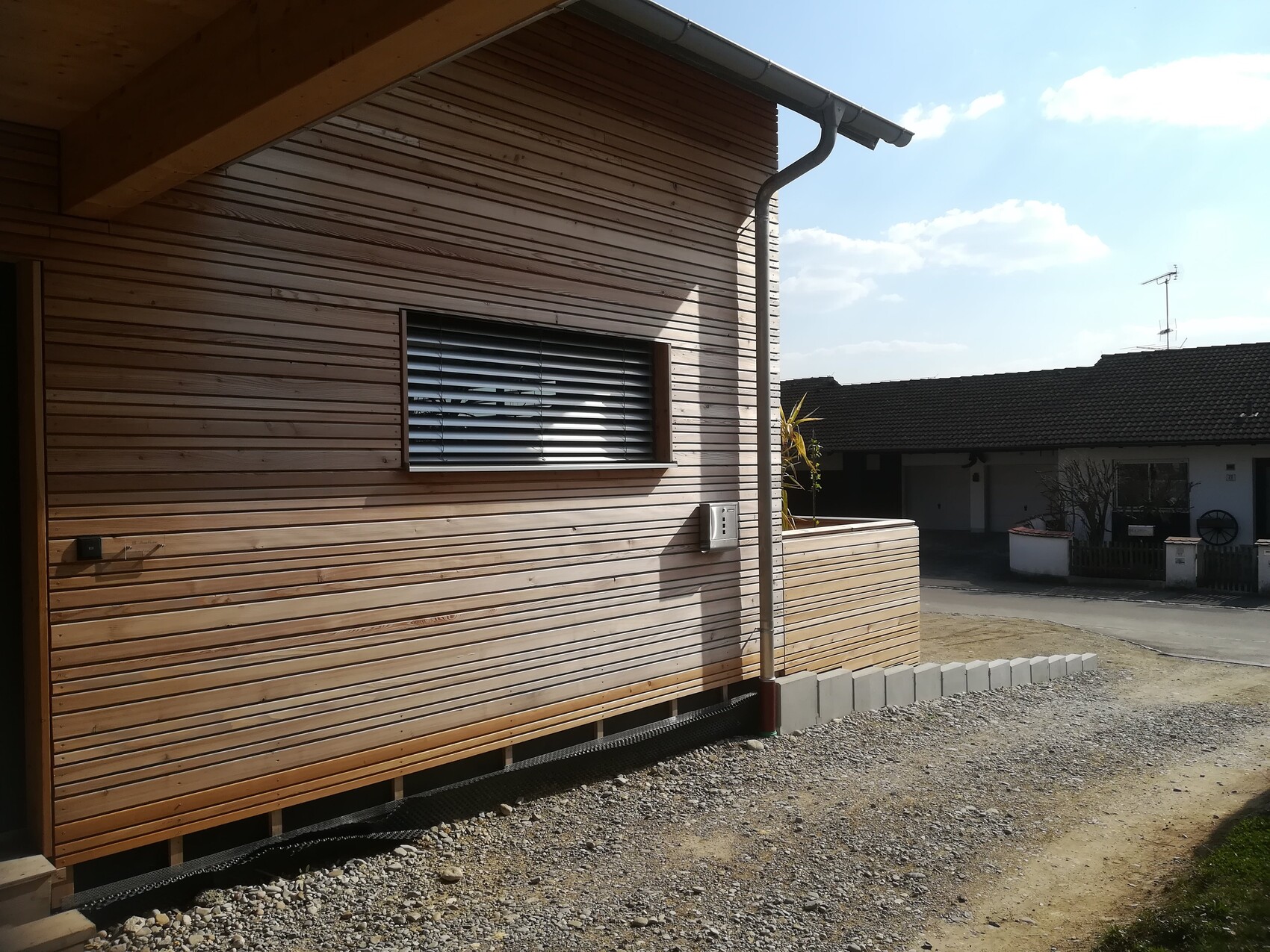 Holzverkleidung der Hausfassade mit Fenster und Jalousien, Regenrinne, Kiesweg vor dem Haus.