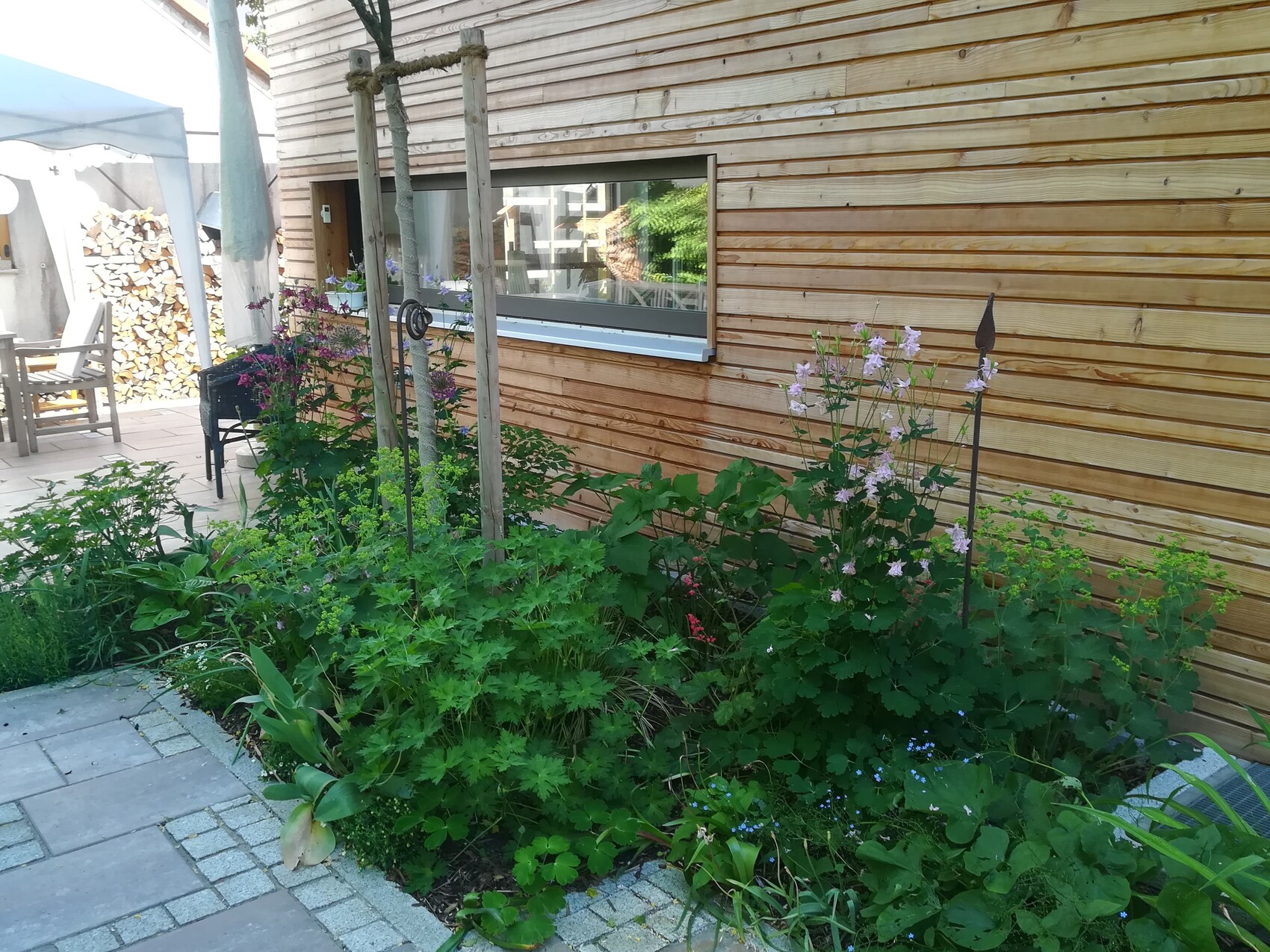 Gartenbeet am Holzhaus mit blühenden Blumen, Steinpfad, Terrasse und Holzstapel im Hintergrund.