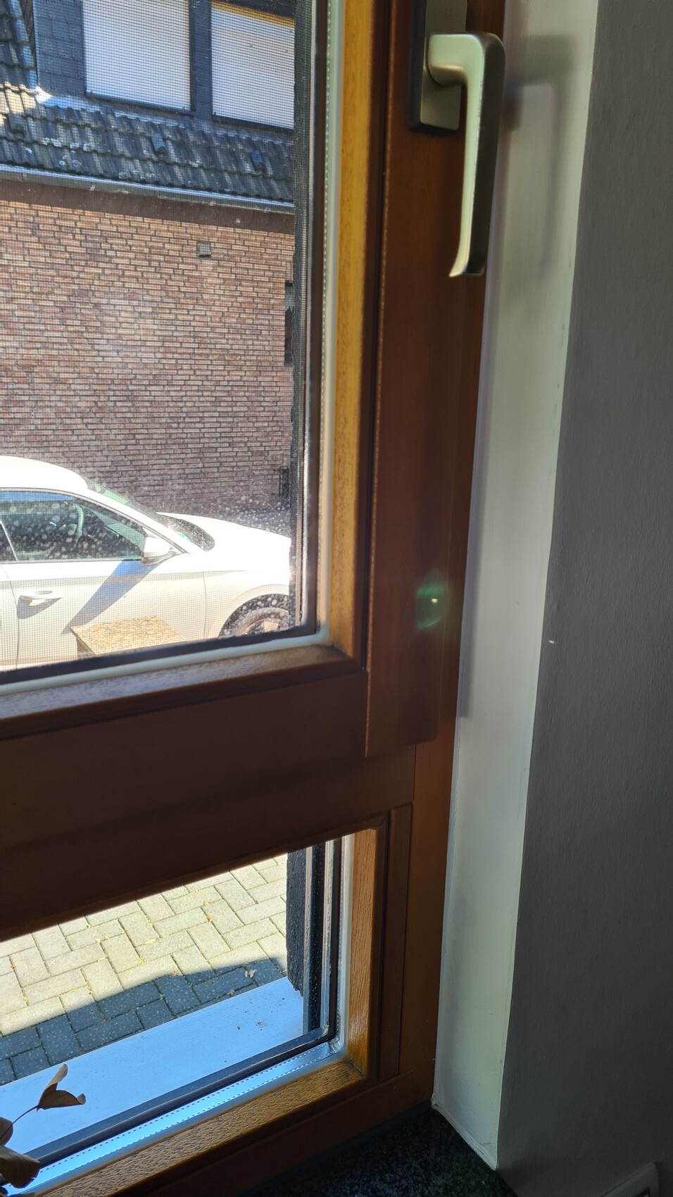 Fenster mit hellem Holzrahmen und Griff; draußen steht ein weißes Auto vor einer Ziegelmauer.