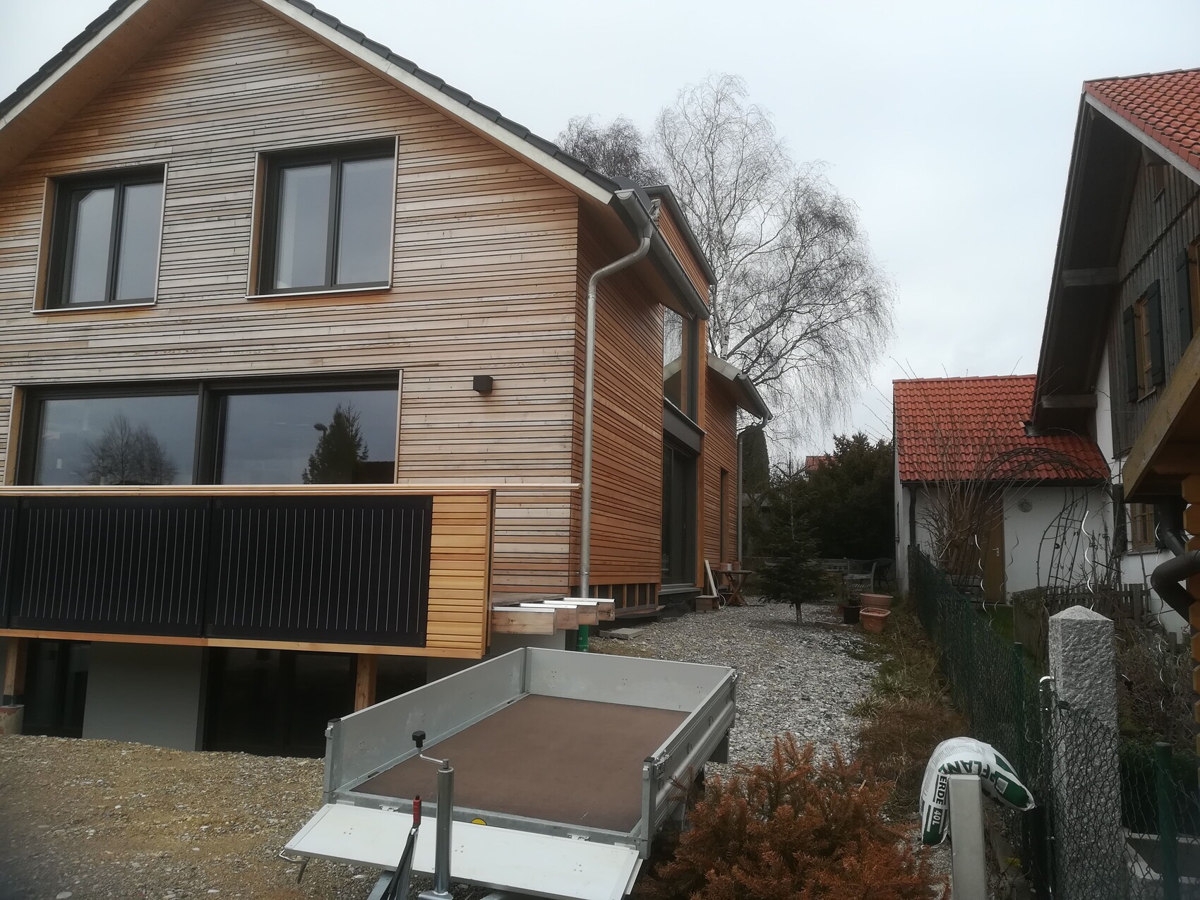 Modernes Holzhaus im Bau mit großer Fensterfront, Holzverkleidung und Anhänger auf Kiesweg.