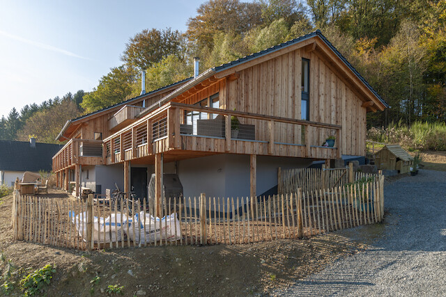 Holzhaus mit überdachter Terrasse und Balkon am Hang, umzäunter Gartenbereich.