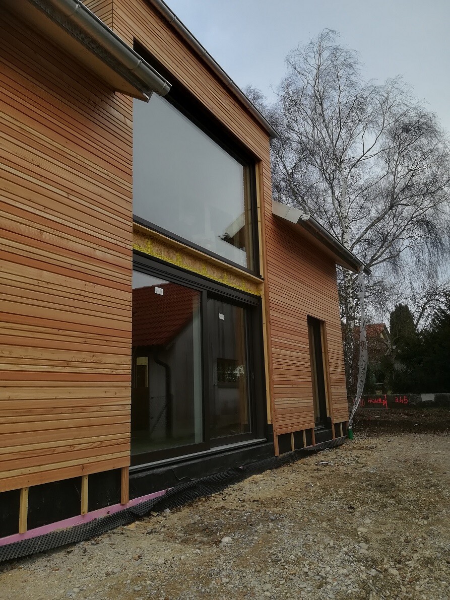 Außenansicht eines Neubauhauses mit Holzwandverkleidung, großen Fenstern und Baugrund.