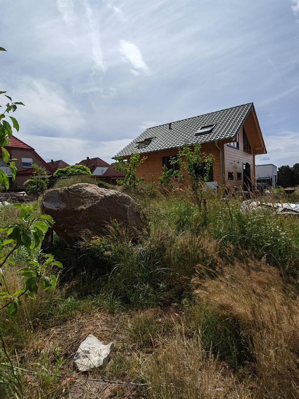 Holzhaus im Bau auf grasigem Grundstück, großer Felsbrocken im Vordergrund, Nachbarhäuser im Hintergrund.