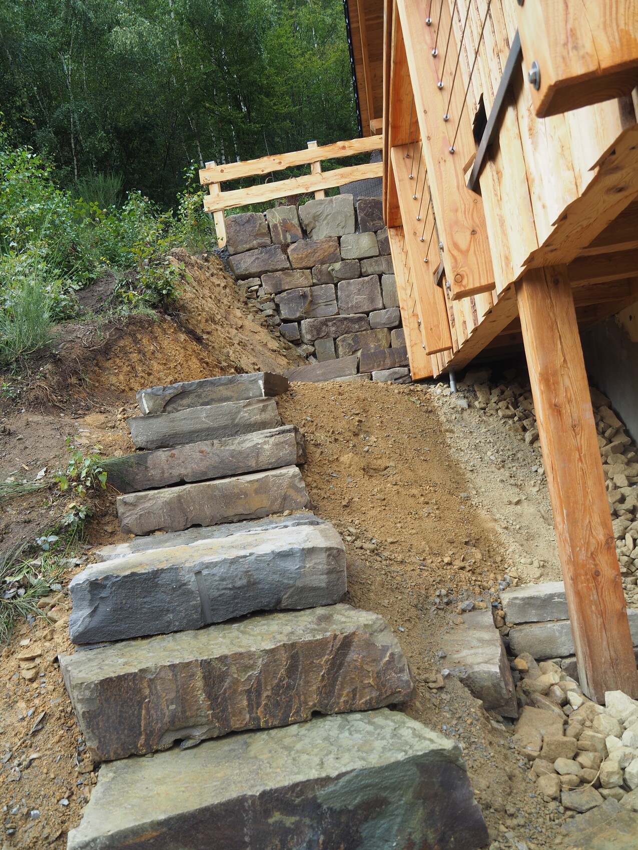 Hangsicherung aus Stein mit Treppen aus großen Blöcken neben Holzhaus, Steinmauer im Hintergrund