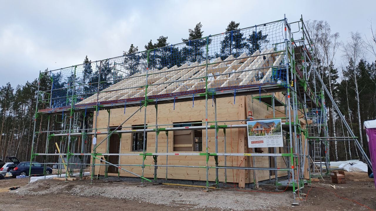 Holzhaus im Bau mit Gerüst, Dachstuhl montiert, Wald im Hintergrund.