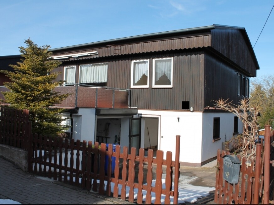 Zweistöckiges Haus mit dunkler Holzfassade und weißem Sockel, offener Garagenbereich, Holzzaun.