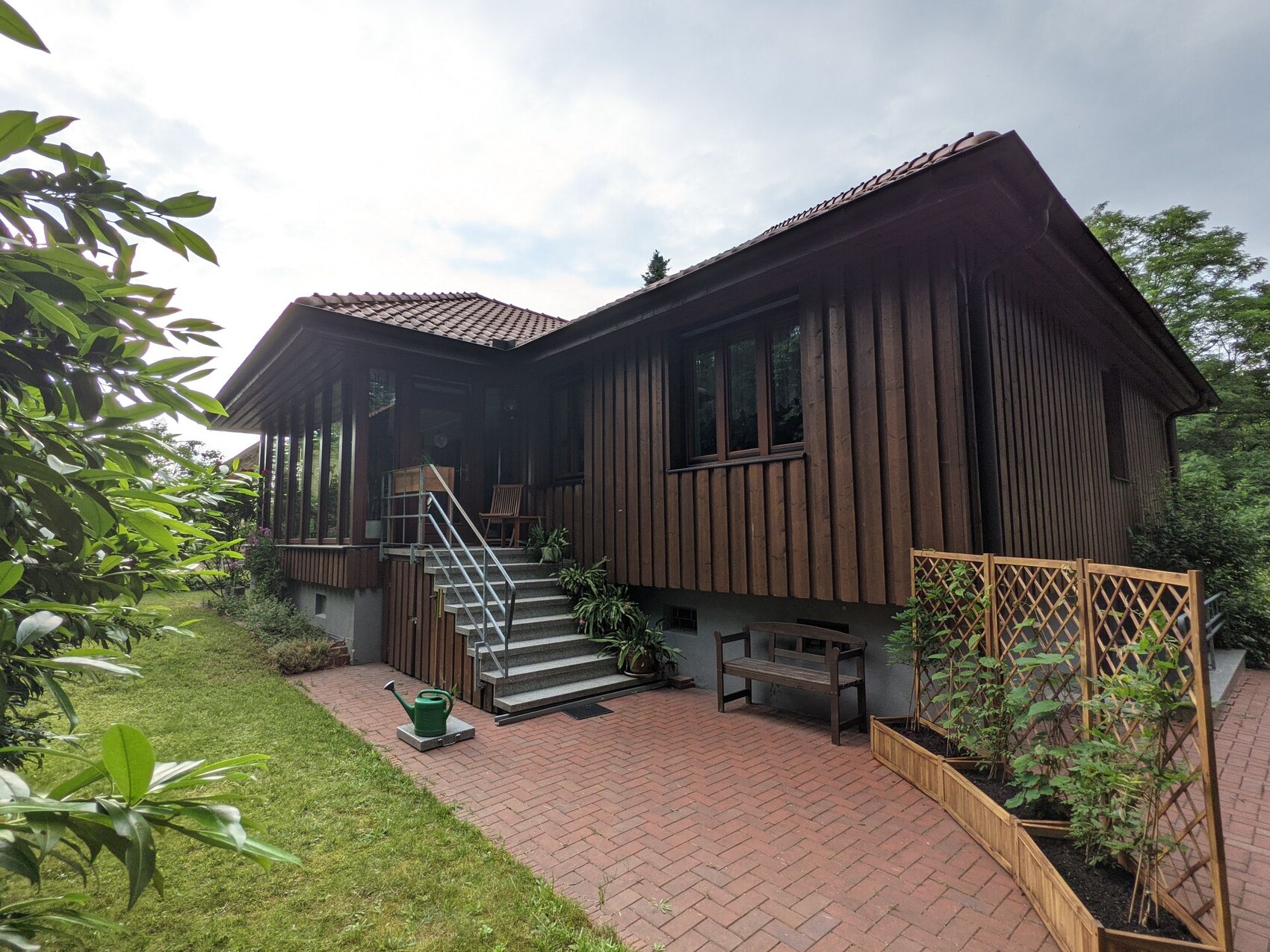 Dunkelbraunes Holzhaus mit Veranda, Treppe, Gartenpflanzen und Pflanzkästen auf Ziegelterrasse.