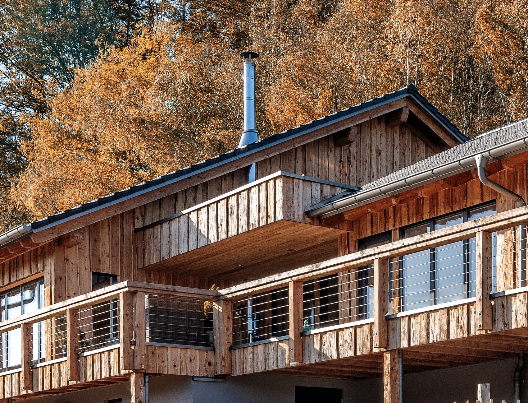 Holzhaus mit Balkon-Veranda aus Holz, Geländer mit Drahtseilen, Herbstlaub dahinter, Kaminrohr