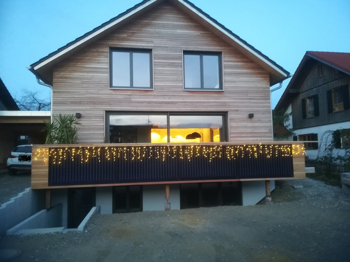 Zweistöckiges Holzhaus mit beleuchteter Terrasse; Lichterketten am Geländer und warmes Innenlicht